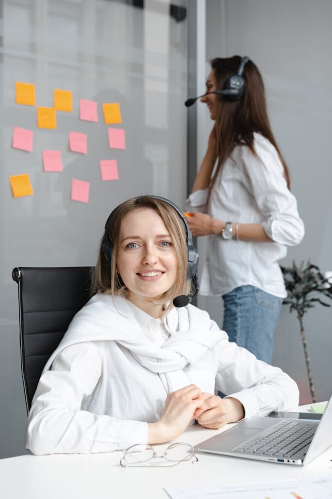 Two female customer support agents smiling and working with laptops and headsets in an office.